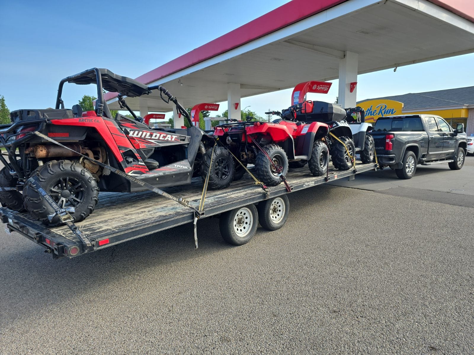 A deck trailer loaded with a side-by-side and three ATVs at a fuel stop